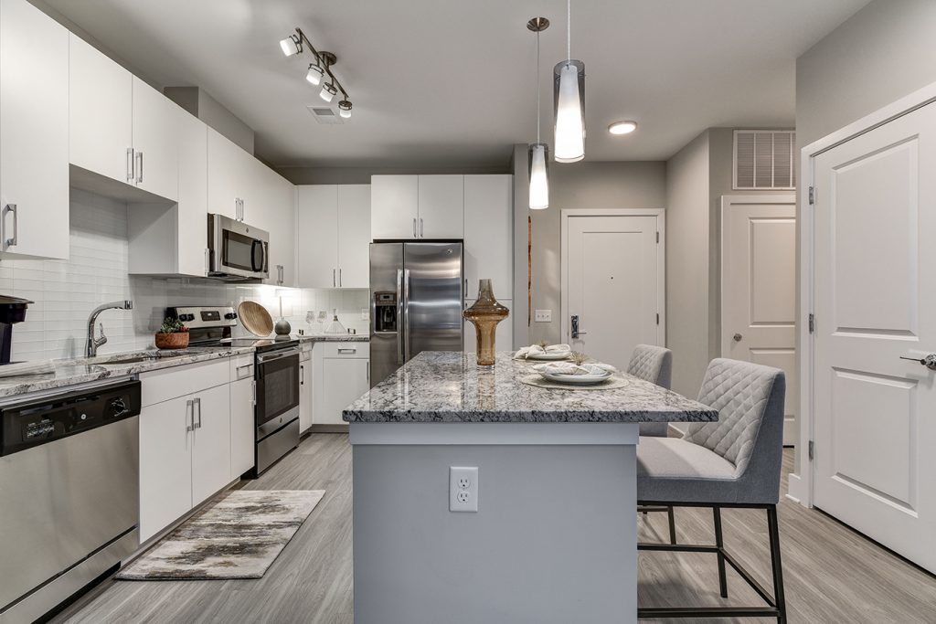 Model apartment kitchen with white cabinets, stainless steel appliances, and large kitchen island with barstool seating and pendant lighting