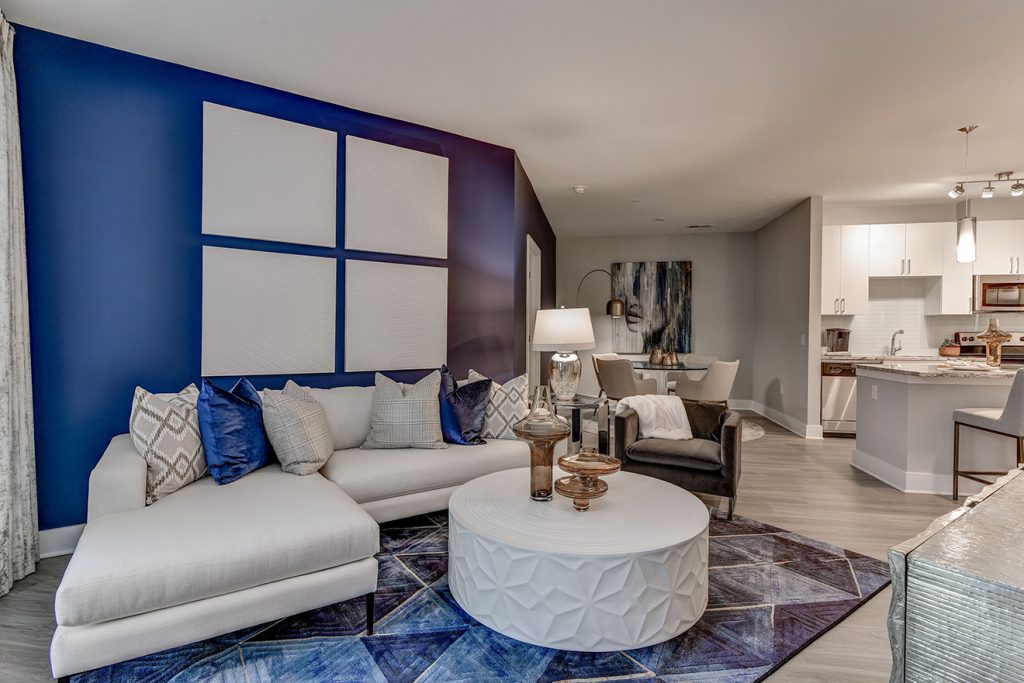 Model apartment living room with blue accent wall, white sectional sofa, wood-style flooring, and view of kitchen